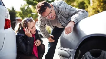 A couple check the damage to the front of their silver car while the woman calls her 州立农场代理.